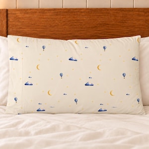 May include: A rectangular pillowcase with a cream-colored background featuring a repeating pattern of small blue hot air balloons, dark blue clouds, yellow crescent moons, and scattered yellow stars across the entire surface.