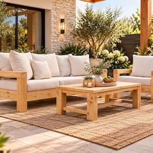 May include: A patio furniture set consisting of a wooden sofa and a matching armchair, both featuring thick off-white cushions. A rectangular wooden coffee table sits in the center, topped with a small woven tray holding decorative items.