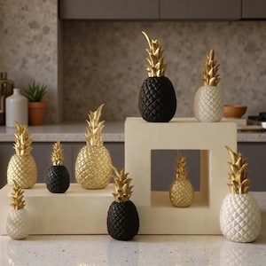 May include: A collection of pineapple-shaped sculptures in various sizes, featuring textured bodies in black, white, and gold finishes with prominent gold-coloured leaves. The sculptures are arranged on and around a light-coloured rectangular display block.