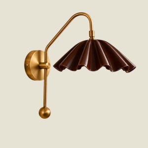 May include: A wall lamp featuring a brass-toned metal arm and a circular wall mount. The arm extends outward and curves to support a dark brown, ruffled, bell-shaped shade. A small spherical brass-toned finial hangs below the arm.