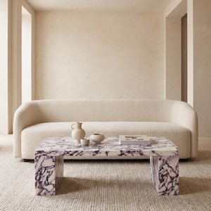 Puede incluir: Una mesa de centro rectangular de mármol blanco con vetas de color púrpura oscuro, con dos patas de losa gruesas. Sobre la mesa hay una pequeña jarra de color crema, un cuenco a juego y un libro cerrado. Detrás hay un sofá curvo color crema.