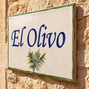 Könnte beinhalten: Ein rechteckiges Keramik-Wandschild mit cremefarbenem Hintergrund und einem dünnen grünen Rand. Der Schriftzug El Olivo ist in blauer Schreibschrift aufgemalt, ergänzt durch eine kleine, detaillierte Abbildung eines grünen Olivenzweigs mit blauen Blüten.