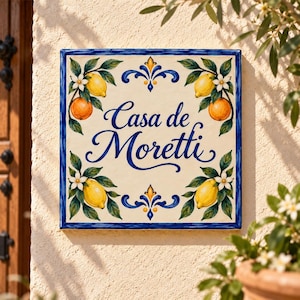 Op de afbeelding: Een vierkante keramische wandplaat met een crèmekleurige achtergrond en een blauwe rand. De tekst 'Casa de Moretti' staat in blauw schrift in het midden. Het ontwerp toont geschilderde gele citroenen, sinaasappels en witte bloesems.