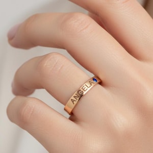 Peut inclure: Une bague anneau dorée portant le nom ANGELA gravé en lettres capitales sur la surface extérieure, agrémentée d'une petite pierre précieuse ronde d'un bleu profond sertie à côté du nom sur l'anneau métallique.