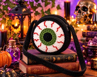 May include: A round, crocheted crossbody bag featuring a large eyeball design with a black pupil, green iris, and white sclera with red bloodshot veins. The bag has a black crocheted strap and a black zipper closure along the top edge.