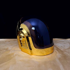 May include: A gold-coloured helmet featuring a smooth, reflective metallic finish and a large, dark, tinted visor that covers the front. The side of the helmet displays intricate, recessed geometric detailing and a circular accent piece.