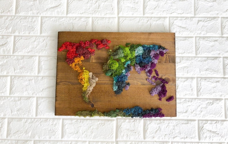 Op de afbeelding: Een wereldkaart gemaakt van geconserveerd mos, waarbij elk continent een andere kleur heeft. De kaart is gemonteerd op een houten plank en heeft een natuurlijke houten afwerking.