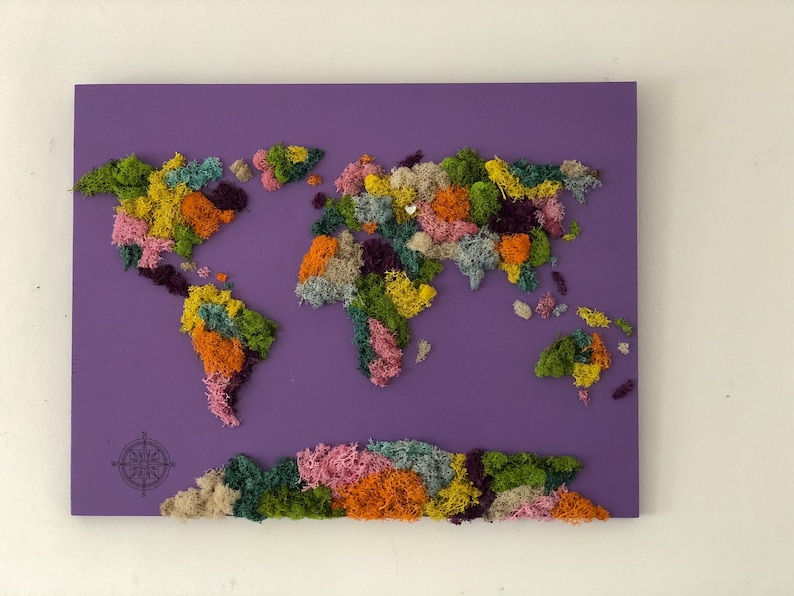 Op de afbeelding: Een wereldkaart gemaakt van geconserveerd mos op een paarse achtergrond. Het mos is gerangschikt in de vorm van de continenten en is in verschillende kleuren, waaronder groen, bruin, roze en oranje. Een kompas bevindt zich in de linkerbenedenhoek.