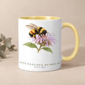 May include: A white ceramic mug with a yellow interior and handle, featuring a detailed illustration of a rusty patched bumble bee on a purple flower. The text RUSTY PATCHED BUMBLE BEE and Bombus affinis is printed below the illustration.
