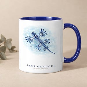 Peut inclure: Un mug en céramique blanche avec un intérieur et une anse bleu foncé. L'avant présente une illustration détaillée d'un glaucus bleu flottant dans l'eau, avec le texte "BLUE GLAUCUS Glaucus atlanticus" imprimé sous l'image.