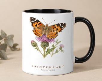 Könnte beinhalten: Ein weißer Keramikbecher mit schwarzem Innenraum und Henkel, verziert mit einer detaillierten Illustration eines orange-schwarzen Distelfalters auf einer violetten Distelblüte, mit dem Text PAINTED LADY Vanessa cardui darunter.