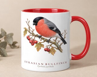 Könnte beinhalten: Ein weißer Keramikbecher mit rotem Innenraum und Henkel, verziert mit einer detaillierten Illustration eines Gimpels auf einem Zweig mit roten Beeren. Der Text EURASIAN BULLFINCH Pyrrhula pyrrhula ist unter dem Vogel aufgedruckt.
