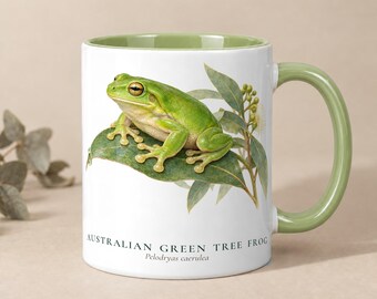 May include: A white ceramic mug with a light green interior and handle, featuring a detailed illustration of a bright green Australian tree frog perched on a leaf. The text reads AUSTRALIAN GREEN TREE FROG and Pelodryas caerulea.