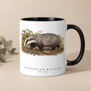 May include: A white ceramic mug with a black interior and handle. The exterior features a detailed illustration of a European badger walking through foliage, with the text "EUROPEAN BADGER" and "Meles meles" printed below the image.