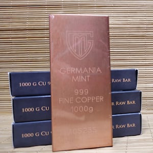 Può includere: Una barra di rame rettangolare con finitura metallica, che presenta il logo Germania Mint, il testo 999 FINE COPPER, 1000g e un numero di serie. È posizionata davanti a una pila di scatole blu scuro etichettate 1000 G CU 999 RAW BAR.