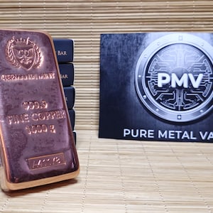Könnte beinhalten: Ein rechteckiger 1000 g Feinkupferbarren der Germania Mint mit geprägtem Schildlogo, dem Text 999.9 FINE COPPER 1000 G und einer Seriennummer. Dahinter befindet sich eine schwarze Karte mit einem silbernen Logo und dem Text PMV PURE METAL VAULT.