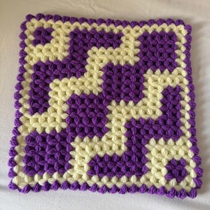 Puede incluir: Un agarrador de cocina cuadrado tejido a ganchillo con un patrón geométrico de puntos puff alternados en color violeta y crema. El diseño consiste en bloques y líneas interconectados, con un borde de puntos puff violetas alrededor.