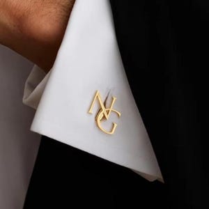 Op de afbeelding: Een goudkleurige manchetknop met de in elkaar grijpende hoofdletters N en G, bevestigd door de knoopsgaten van een wit overhemdmanchet, dat gedeeltelijk zichtbaar is tegen een effen zwarte stoffen achtergrond.