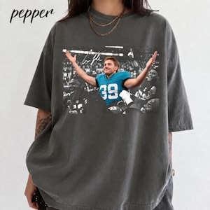 May include: A charcoal gray short-sleeve t-shirt featuring a graphic print of a football player wearing a teal jersey with the number 39, arms raised in celebration, with the word pepper printed in the upper left corner.