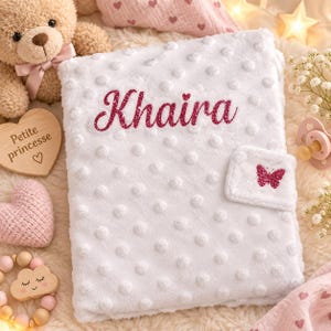 Peut inclure: Une couverture en peluche blanche avec une texture à pois en relief, présentant le prénom Khaira en écriture cursive rose foncé pailletée et une petite applique papillon rose foncé pailletée sur la patte d'angle, entourée d'accessoires.