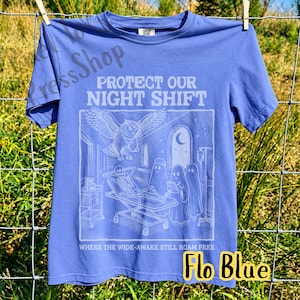 May include: A periwinkle blue short-sleeve t-shirt featuring a white graphic print. The text reads PROTECT OUR NIGHT SHIFT and WHERE THE WIDE-AWAKE STILL ROAM FREE. The illustration depicts an owl, a skeleton on a gurney, and three ghosts.