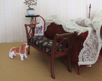 May include: A small, brown wooden bench with a floral patterned cushion and a red pillow with a cross-stitch design. A small, white ceramic planter with a miniature tree sits on a table behind the bench. A brown and white dog figurine stands in front of the bench.