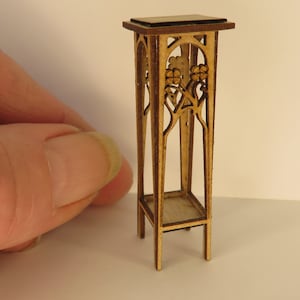 Puede incluir: Un soporte de madera en miniatura con una parte superior de color marrón oscuro y tallas intrincadas. El soporte tiene un estante y está hecho de madera marrón claro.