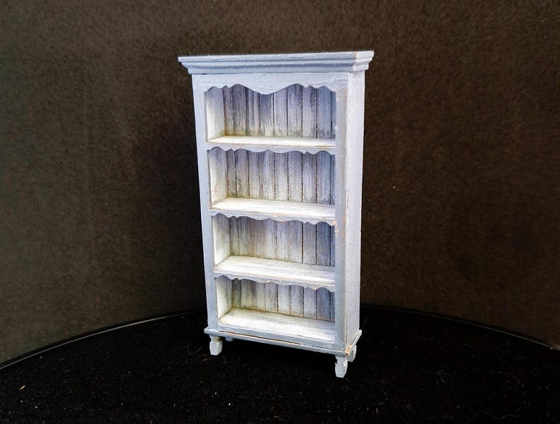 May include: A white painted wooden bookshelf with four shelves and decorative scalloped edges. The shelves are made of wood planks with a distressed finish.