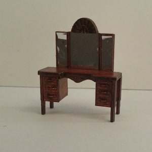 Op de afbeelding: Een miniatuur houten kaptafelset met een driedelige spiegel en laden. De kaptafel is in donkerbruin geverfd en heeft een decoratief ontwerp aan de voorkant.