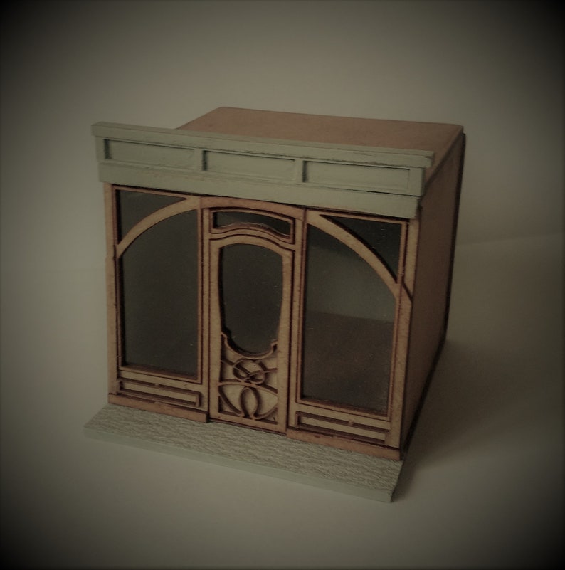May include: A miniature wooden model of a storefront with a gray roof and a glass door with a decorative design. The storefront has a wooden frame and a small ledge in front.