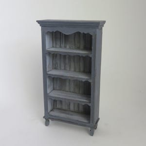 May include: A gray wooden bookcase with four shelves and a decorative top. The shelves have a scalloped edge and the back of the bookcase is made of wood slats.