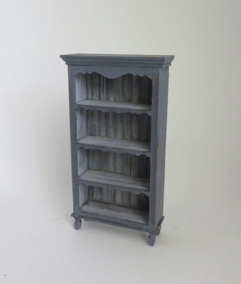 May include: A gray wooden bookcase with four shelves and a decorative top. The shelves have a scalloped edge and the back of the bookcase is made of wood slats.