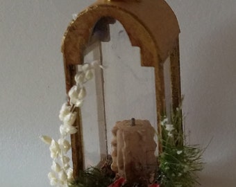 May include: A gold-colored lantern with a clear glass window and a white candle inside. The lantern is decorated with white flowers and green foliage.