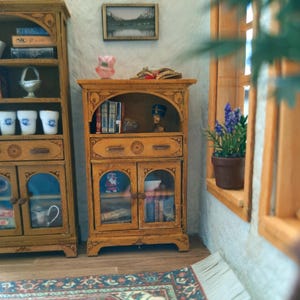 May include: A miniature wooden cabinet with two glass-paneled doors, a drawer, and an open shelf containing books and a small bust. The cabinet is finished in a light brown tone and features carved details on the drawer and frame edges.