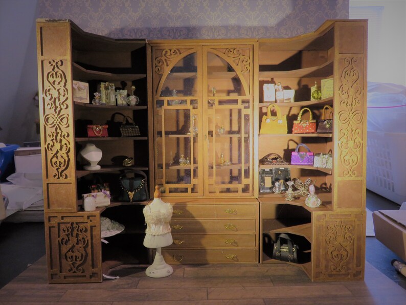 May include: A wooden cabinet with glass doors and shelves, filled with decorative items, including purses, bottles, and jewelry. The cabinet has intricate carvings and a distressed finish.