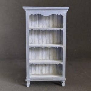 May include: A gray wooden bookshelf with four shelves and a decorative top. The shelves have a scalloped edge and the bookshelf has turned legs.