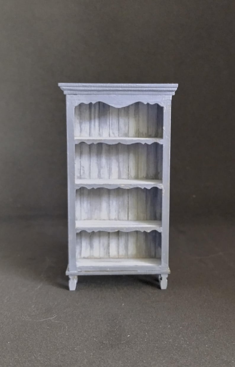 May include: A gray wooden bookshelf with four shelves and a decorative top. The shelves have a scalloped edge and the bookshelf has turned legs.