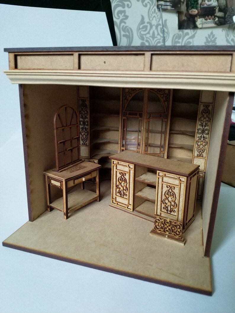 May include: A miniature wooden model of a shop interior with shelves, a counter, and a small table with a mirror. The model is made of light brown wood and has intricate details.