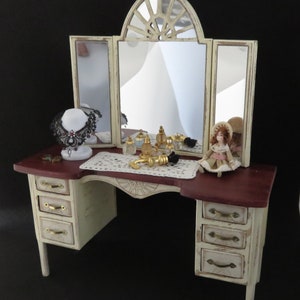 May include: A white vanity with a three-panel mirror, a burgundy top, and gold hardware. The vanity has three drawers on each side and a lace doily on the top. There are several bottles of perfume and a gold faucet on the vanity. A doll wearing a white dress with pink accents is sitting on the vanity.