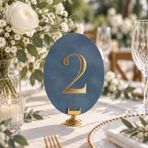 May include: An oval-shaped table number sign in a dusty blue color featuring a large, metallic gold numeral two in the center. The sign is held upright by a small, circular gold metal stand, creating a clear display for event seating.