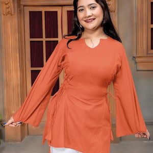 May include: A burnt orange long-sleeved tunic featuring a notched neckline and side lace-up details with hanging tassels, paired with white trousers. The tunic has wide, flared sleeves and a straight hemline that falls below the waist.