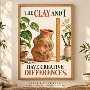 May include: A framed wall art print featuring the text THE CLAY AND I HAVE CREATIVE DIFFERENCES. The illustration shows a terracotta-colored vase with pink flowers, a wooden ruler, a potted plant, and paintbrushes in a jar.