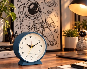 May include: A round, matte blue analog quartz desk clock with a white face, black numerals, black hour and minute hands, and a thin yellow second hand. The clock features a sturdy base and is positioned on a wooden surface.
