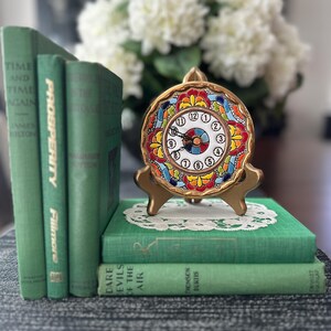 May include: A decorative gold-toned clock with a colorful floral design, resting on a stack of green books. The clock face displays Roman numerals. A bouquet of white flowers is in the background. The books are titled "Time and Time Again" and "Dare Evils of the Air."
