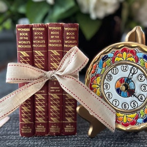 May include: Four small red books with gold lettering titled "Wonder Book of the World's Progress" are stacked and tied together with a beige ribbon. The books are sitting on a gray textured surface next to a gold clock with a colorful painted face.