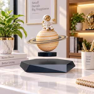 Könnte beinhalten: Ein schwebendes Saturn-Ornament mit einer weiß-goldenen Astronautenfigur, die obenauf sitzt. Der Planet hat braune und beige Bänder mit einem Ringsystem und schwebt über einem schwarzen, sechseckigen Sockel.