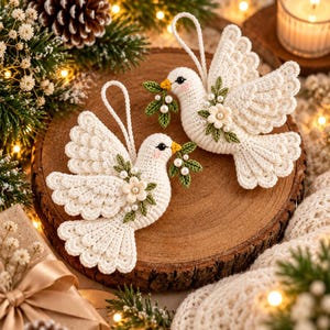 May include: Two cream-colored crocheted dove ornaments, each featuring a small yellow beak, a black eye, and a green leafy sprig with white beads held in its beak. Each dove is adorned with a small white flower and green leaves.