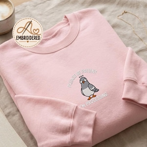 May include: A light pink crewneck sweatshirt featuring an embroidered graphic of a grey pigeon with wide eyes. The text above and below the bird reads, "PLEASE BE PATIENT I'M PROCESSING" in white capital letters. The garment has long sleeves.