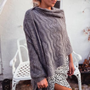 May include: A grey knitted poncho featuring a textured wavy cable pattern and ribbed edges. The garment has a loose, draped fit with a wide neckline and long sleeves, showcasing intricate stitch details throughout the fabric.