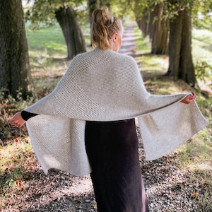 May include: A large, rectangular, light beige knit shawl featuring a combination of textured stitch patterns, including a honeycomb-like design on one side and a ribbed knit pattern on the other, draped horizontally across the back.